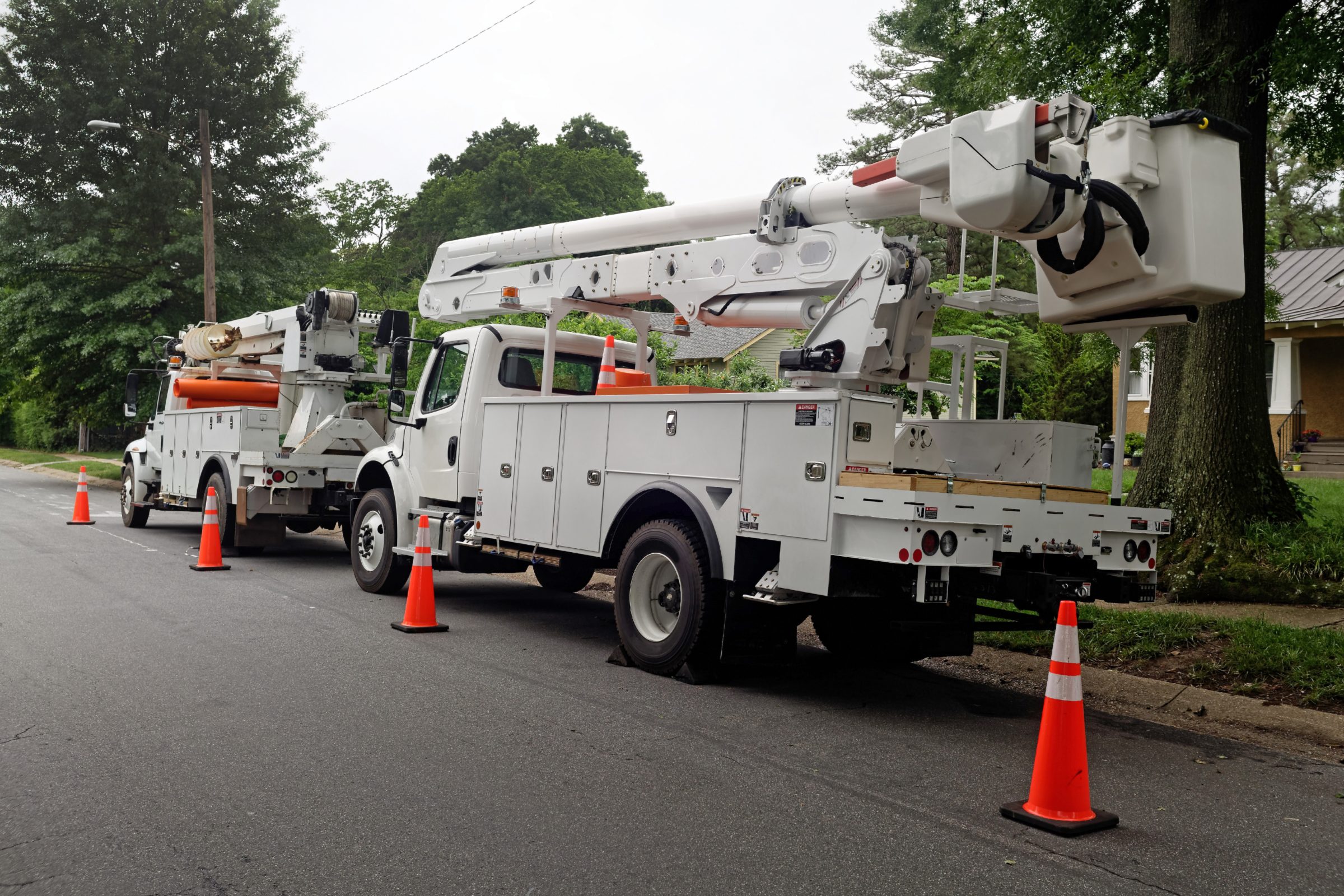 Utility bucket trucks parked on a residential street behind safety cones